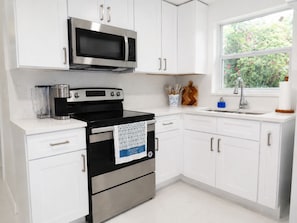 Una cocina con armarios blancos, estufa negra, microondas, cafetera y ventana con vistas a follaje.
