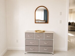 Un mur blanc avec un miroir, une commode grise avec des tiroirs et un rideau bleu.