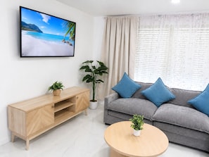 A modern living room with a flat-screen TV, a grey sofa with blue cushions, a wooden coffee table, a wooden TV stand, and a large window with sheer curtains.