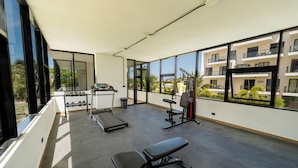 A gym with treadmills, a bench, and weight machines, large windows, and a view of a building with balconies. - Modern Studio Downtown Pool & Lake View + Gym Central Park (Punta Cana)