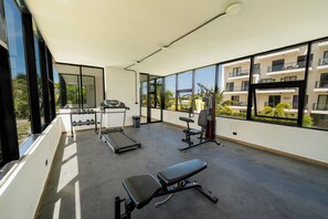 A gym with treadmills, a bench, and weight machines, large windows, and a view of a building with balconies. - Modern Studio Downtown Pool & Lake View + Gym Central Park (Punta Cana)