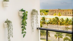 A balcony with hanging plants, a wooden chair, and a view of a hotel and beach. - Modern Studio Downtown Pool & Lake View + Gym Central Park (Punta Cana)