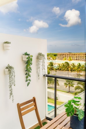 A balcony with hanging plants, a wooden chair, and a view of a hotel and beach. - Modern Studio Downtown Pool & Lake View + Gym Central Park (Punta Cana)