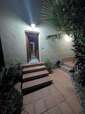 Una escalera conduce a una habitación con una lámpara en el techo, rodeada de plantas en macetas y un muro de piedra.