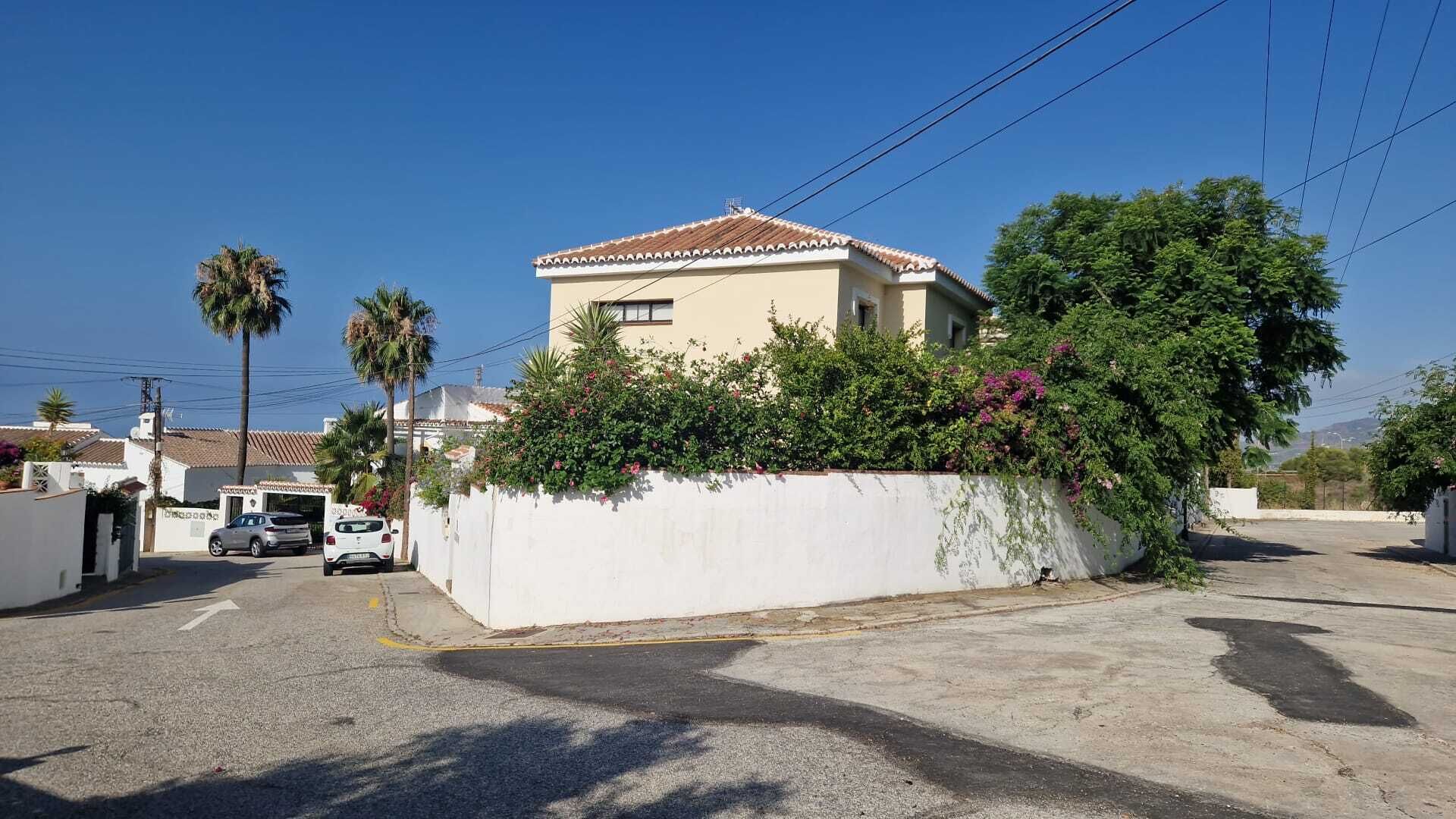 A residential building with a tiled roof, surrounded by a white wall and lush greenery.