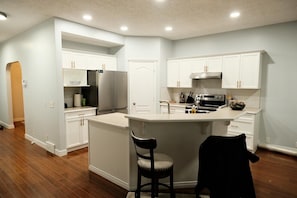 A modern kitchen with white cabinetry, a stainless steel refrigerator, an island with bar stools, and a built-in oven. - Spacious 3 Bedroom Home in McKenzie Towne (Calgary)