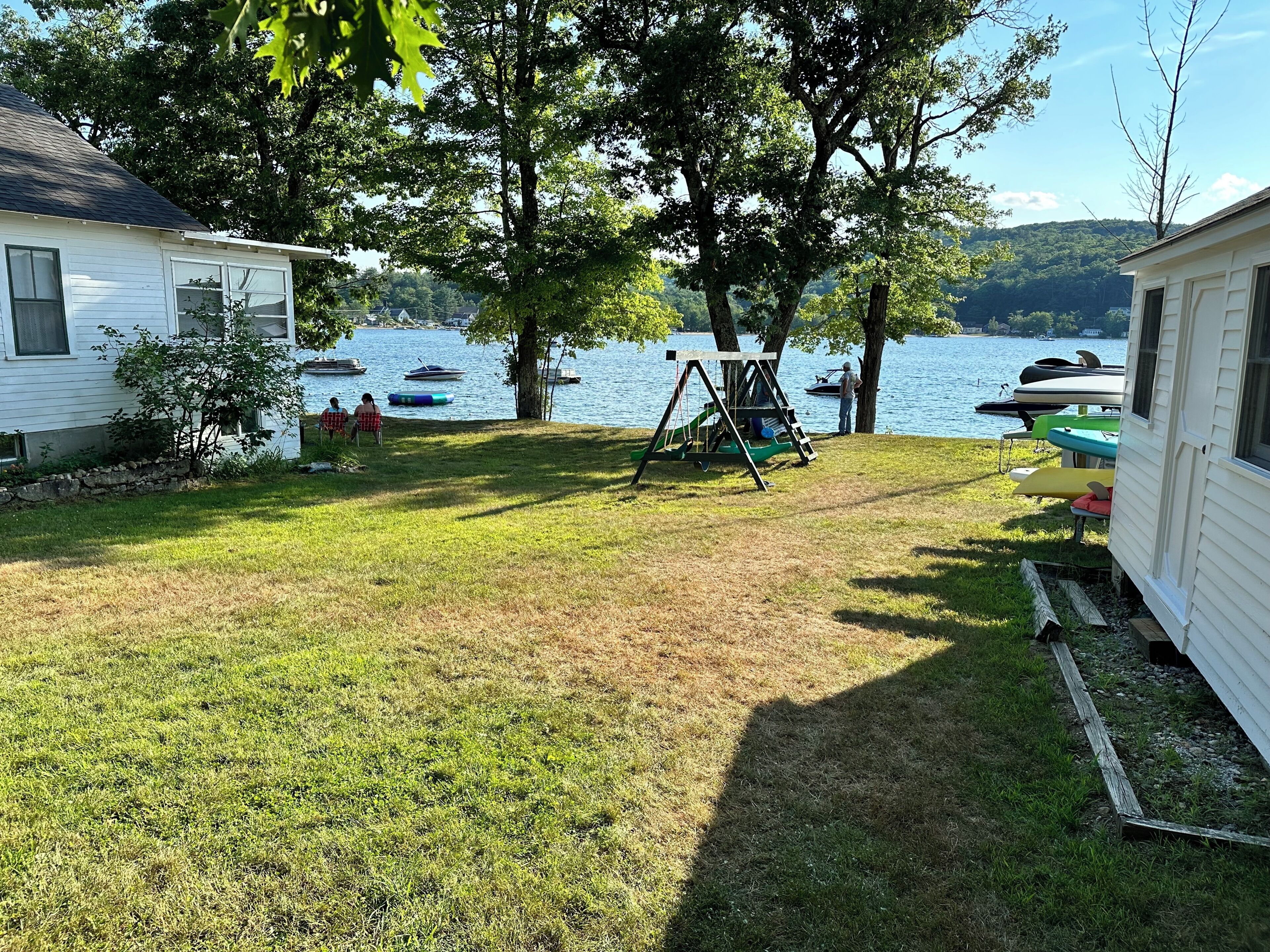 A grassy yard with a swing set, a small tree, and a lake with boats.