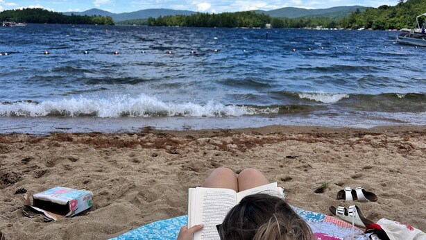 Escena de playa con una persona leyendo un libro, un bolso y un par de sandalias sobre la arena.