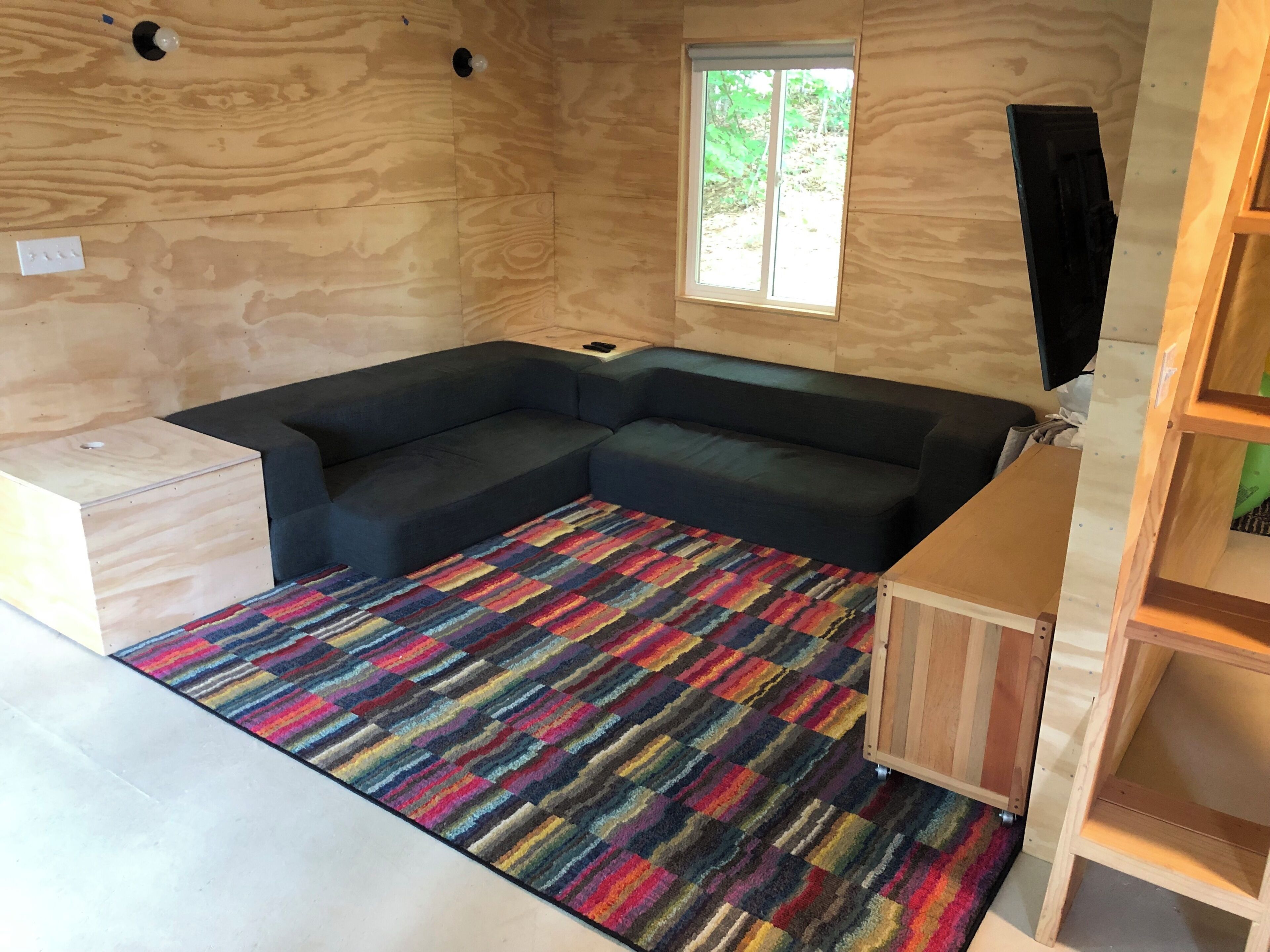 Ein Zimmer mit einer Eckcouch, einem farbenfrohen Streifen Teppich, einem Fenster mit Blick auf Grünflächen und einer Holzkabine.