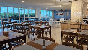 A restaurant with multiple tables and chairs, a counter with a menu, and large windows offering a view of the outdoors. - Beachfront apartment with ocean view in Recreio. (Rio de Janeiro)