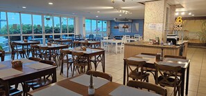 A restaurant with multiple tables and chairs, a counter with a menu, and large windows offering a view of the outdoors. - Beachfront apartment with ocean view in Recreio. (Rio de Janeiro)