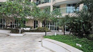 A courtyard with a tiled floor, surrounded by planters with various plants, and a building with multiple balconies and large windows. - Beachfront apartment with ocean view in Recreio. (Rio de Janeiro)