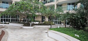A courtyard with a tiled floor, surrounded by planters with various plants, and a building with multiple balconies and large windows. - Beachfront apartment with ocean view in Recreio. (Rio de Janeiro)