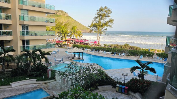 A hotel pool area with a view of the ocean, palm trees, and a beachside bar. - Beachfront apartment with ocean view in Recreio. (Rio de Janeiro)