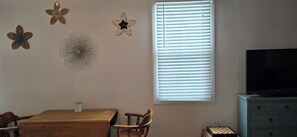 Ein Zimmer mit einem Holztisch, zwei Stühlen, einem Fernseher auf einer Kommode und einem Fenster mit Jalousien.