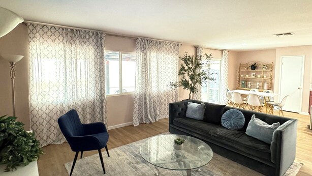 A modern living room with a dark grey sofa, two blue armchairs, a glass coffee table, and a dining area in the background.