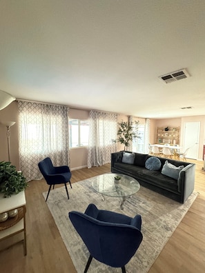 Una sala de estar moderna con un sofá gris oscuro, dos sillones azules, una mesa de centro de vidrio y un comedor al fondo.