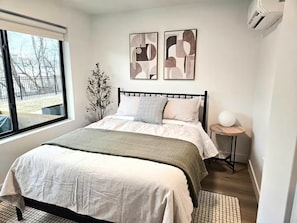Ein Schlafzimmer mit einem Bett, einem Fenster, einer Pflanze, abstrakter Wandkunst und einem Nachttisch mit Lampe.