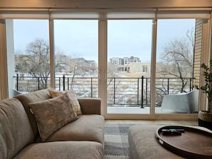 Ein Wohnzimmer mit großem Fenster, einer Couch, einem Couchtisch und Blick auf Gebäude und Bäume.