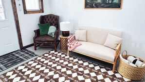 Une pièce avec un tapis à carreaux marron et blanc, un fauteuil en cuir marron avec un coussin vert, un canapé blanc avec un plaid à carreaux, un tableau représentant une forêt et un panier en osier.