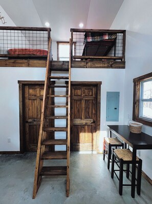 Una habitación tipo loft con una escalera de madera que sube a una cama en zona superior, una mesa de comedor con sillas y un ventanal con vistas.