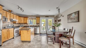 Een keuken met houten kasten, roestvrijstalen apparatuur, een eettafel met stoelen en uitzicht door een glazen deur.