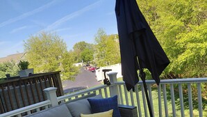 Une terrasse dotée d’un canapé d’angle gris, d’un parasol bleu et offrant une vue sur un parking bordé d’arbres.