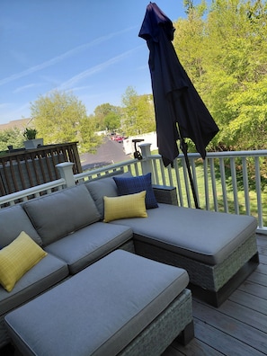 A deck with a gray sectional sofa, a blue umbrella, and a view of a parking area with trees.