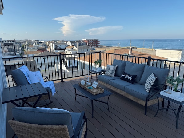 A deck with a sofa, chairs, and a table, overlooking a residential area and the ocean.