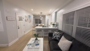 A modern kitchen with a central island, a dining area with bar stools, a living room with a sofa and decorative pillows, and a hallway leading to a bedroom. - New Luxury 3 bedroom 3.5 baths Condo located in the heart of Salisbury Beach. (Salisbury)