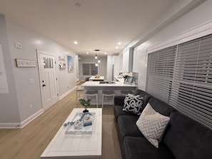 A modern kitchen with a central island, a dining area with bar stools, a living room with a sofa and decorative pillows, and a hallway leading to a bedroom.