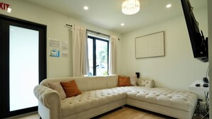 A beige sectional sofa with orange cushions, a large window with curtains, a framed artwork on the wall, and a flat-screen TV on a stand. - World Cup Oasis. 12 min from Beach & Casino. 18 min from World Cup stadium. (Hollywood)