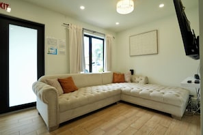Ein beiges Ecksofa mit orangefarbenen Kissen, ein großes Fenster mit Vorhängen, ein gerahmtes Kunstwerk an der Wand und ein Flachbildfernseher auf einem Standfuß.