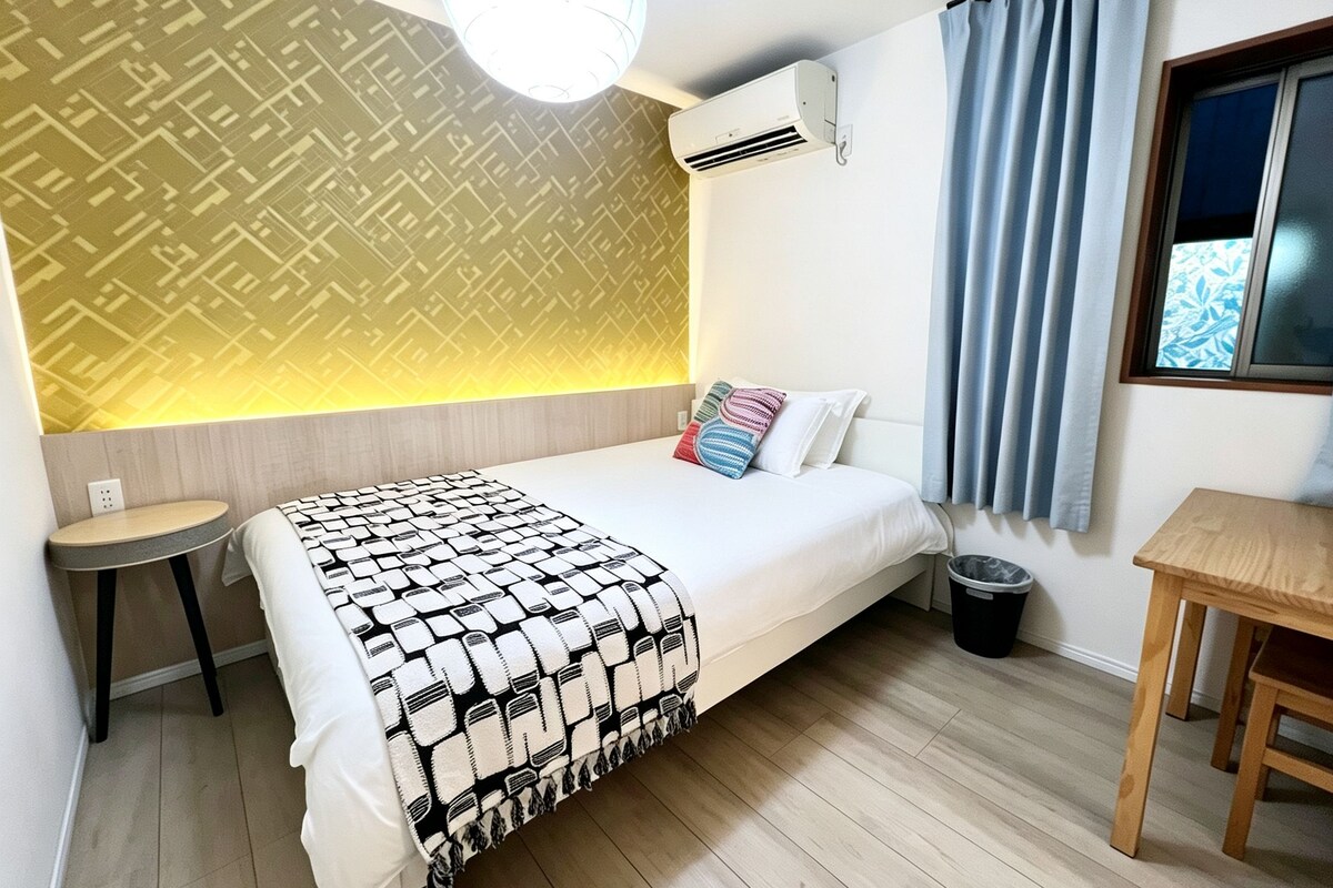 A hotel room with a bed, a small table, a trash bin, and a window with curtains.