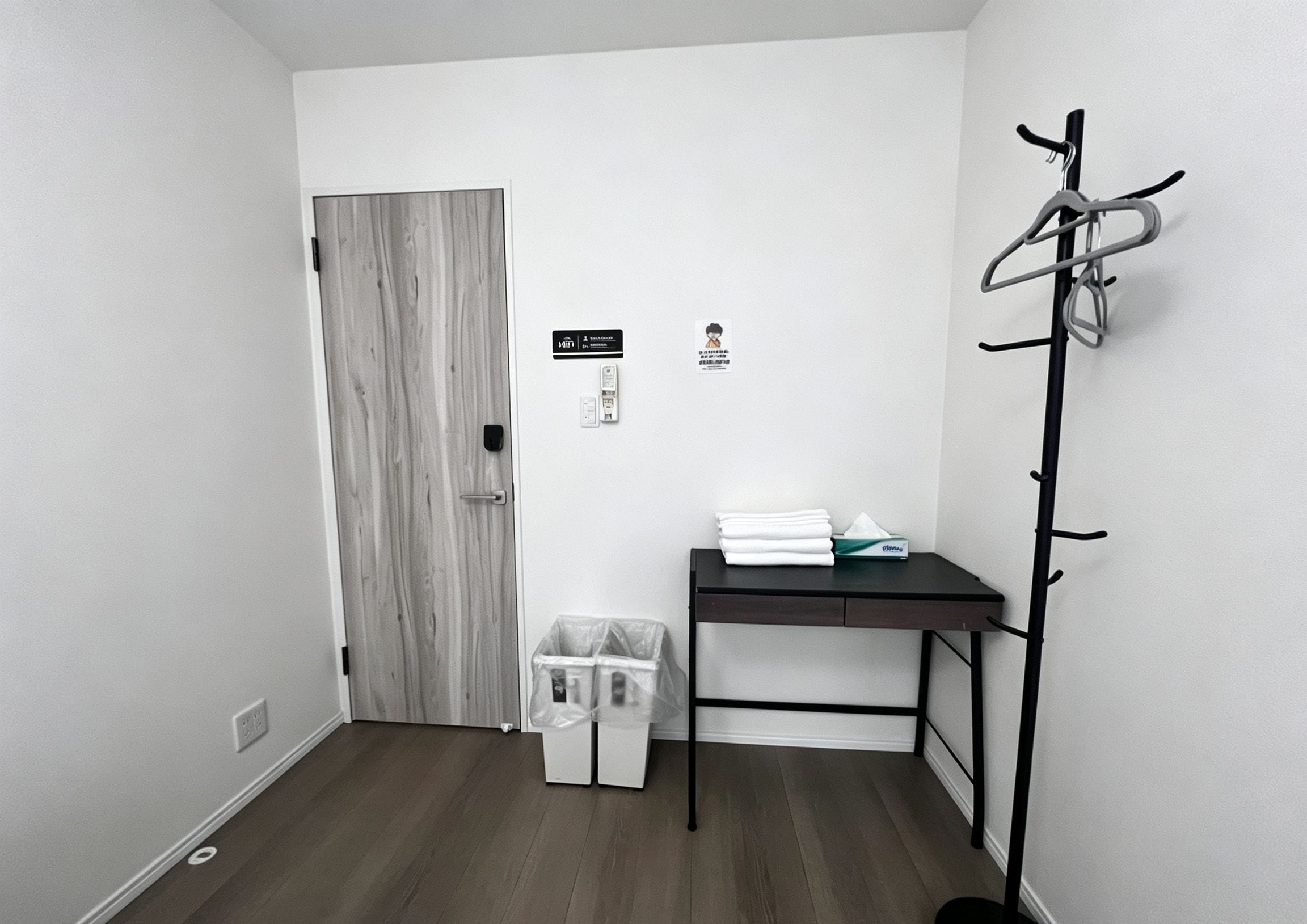 Una habitación con puerta de madera, perchero, pequeña mesa con toallas dobladas y un cesto de basura.