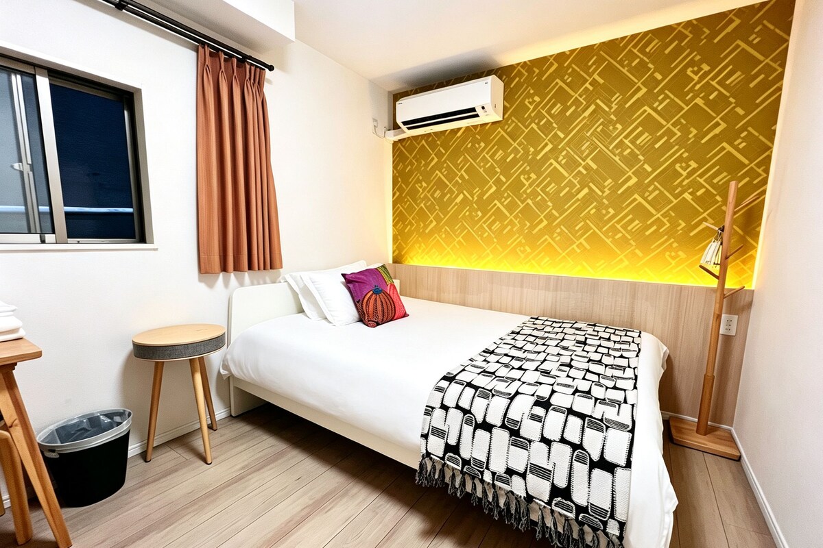 A hotel room with a bed, a small table, a trash bin, an air conditioning unit, and a patterned wall.