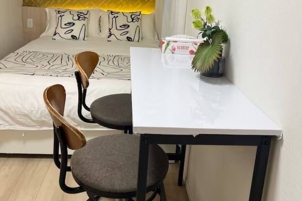 Quarto pequeno e moderno com cama, duas cadeiras, uma mesa e um vaso de planta.
