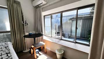 A compact room with a bed, a small table, a chair, a window with a view, and an air conditioning unit.