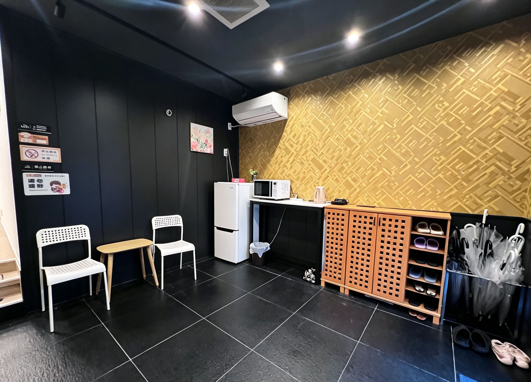 A modern interior with a black and gold patterned wall, a white refrigerator, a microwave, a small table, two white chairs, and a shoe rack.
