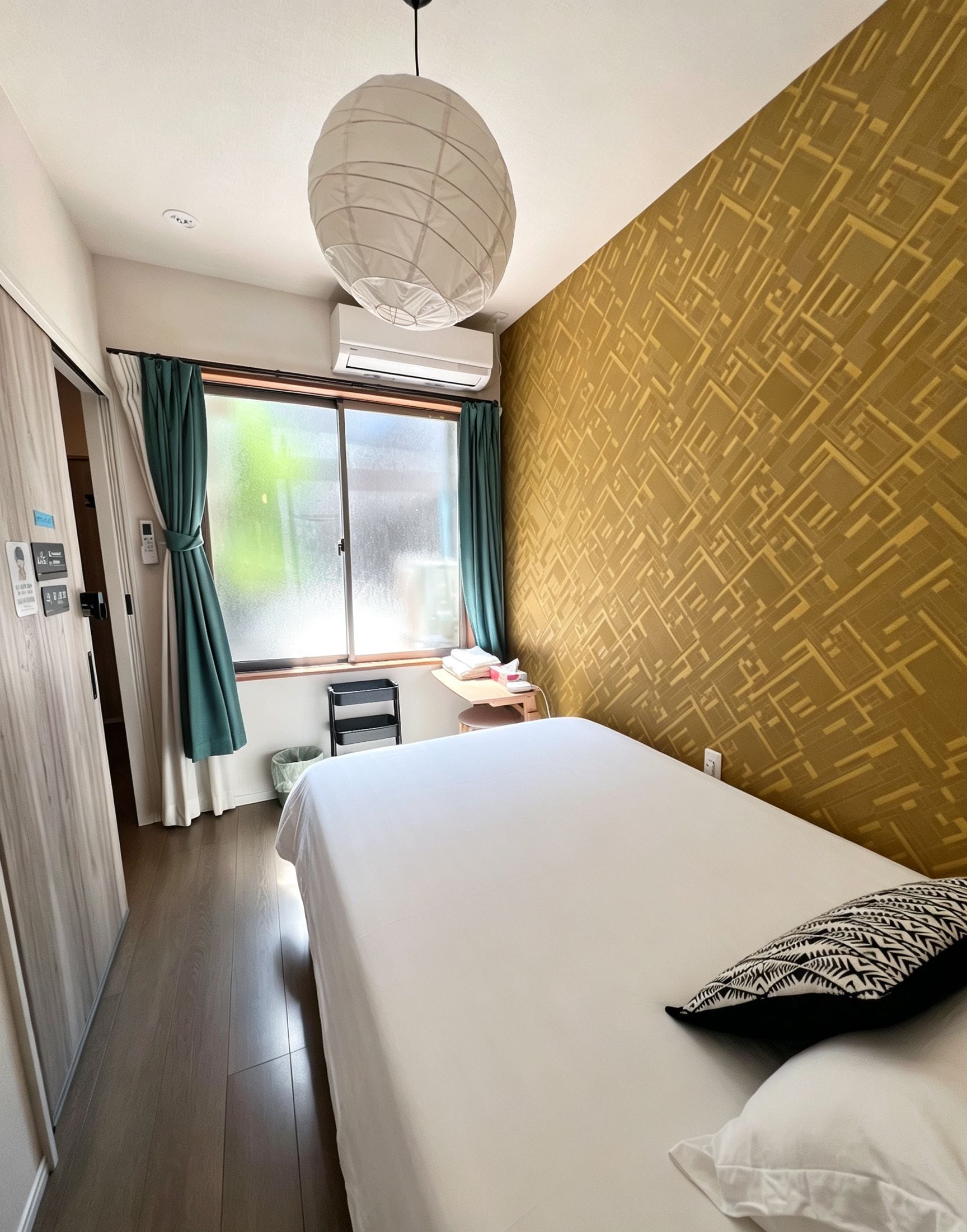 A hotel room with a bed, a window with curtains, an air conditioning unit, a wall with a geometric pattern, and a door with a towel rack.
