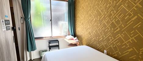 A hotel room with a bed, a window with curtains, an air conditioning unit, a wall with a geometric pattern, and a door with a towel rack.