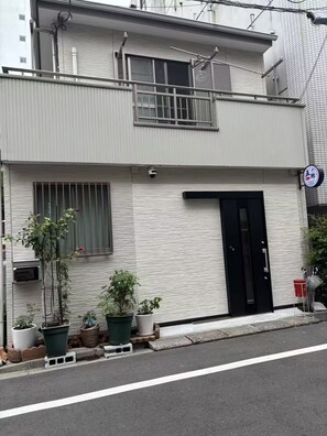 Un edificio moderno de dos pisos, con puerta negra, plantas en macetas y una cámara de seguridad.