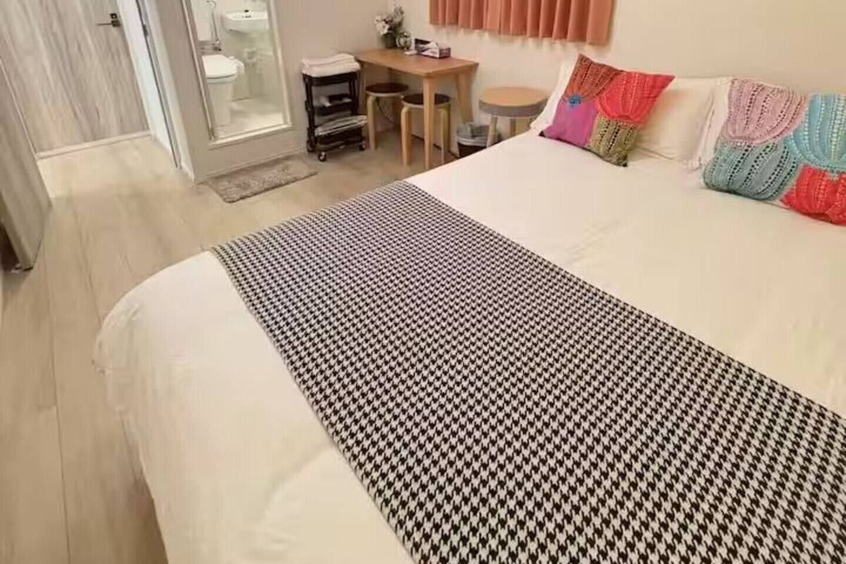 Quarto com cama, escrivaninha, cadeira, luminária pendente e banheiro visível por uma porta aberta.