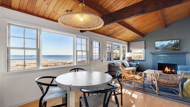 A room with a wooden ceiling, large windows overlooking the beach, a round table with chairs, and a fireplace. - Bay Views, Steps to Beach, Minutes to P-Town (Wellfleet)