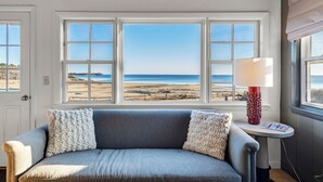 A room with a gray sofa, two white pillows, a red patterned lamp, and a view of the beach through three windows. - Bay Views, Steps to Beach, Minutes to P-Town (Wellfleet)