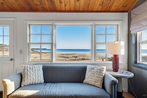 A room with a gray sofa, two white pillows, a red patterned lamp, and a view of the beach through three windows. - Bay Views, Steps to Beach, Minutes to P-Town (Wellfleet)