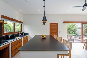 A modern kitchen with a central island, wooden cabinets, a black countertop, a hanging pendant light, and a view of an outdoor deck. - High End Luxury 3BR Villa Walking Distance to Nuanu City (Tabanan)