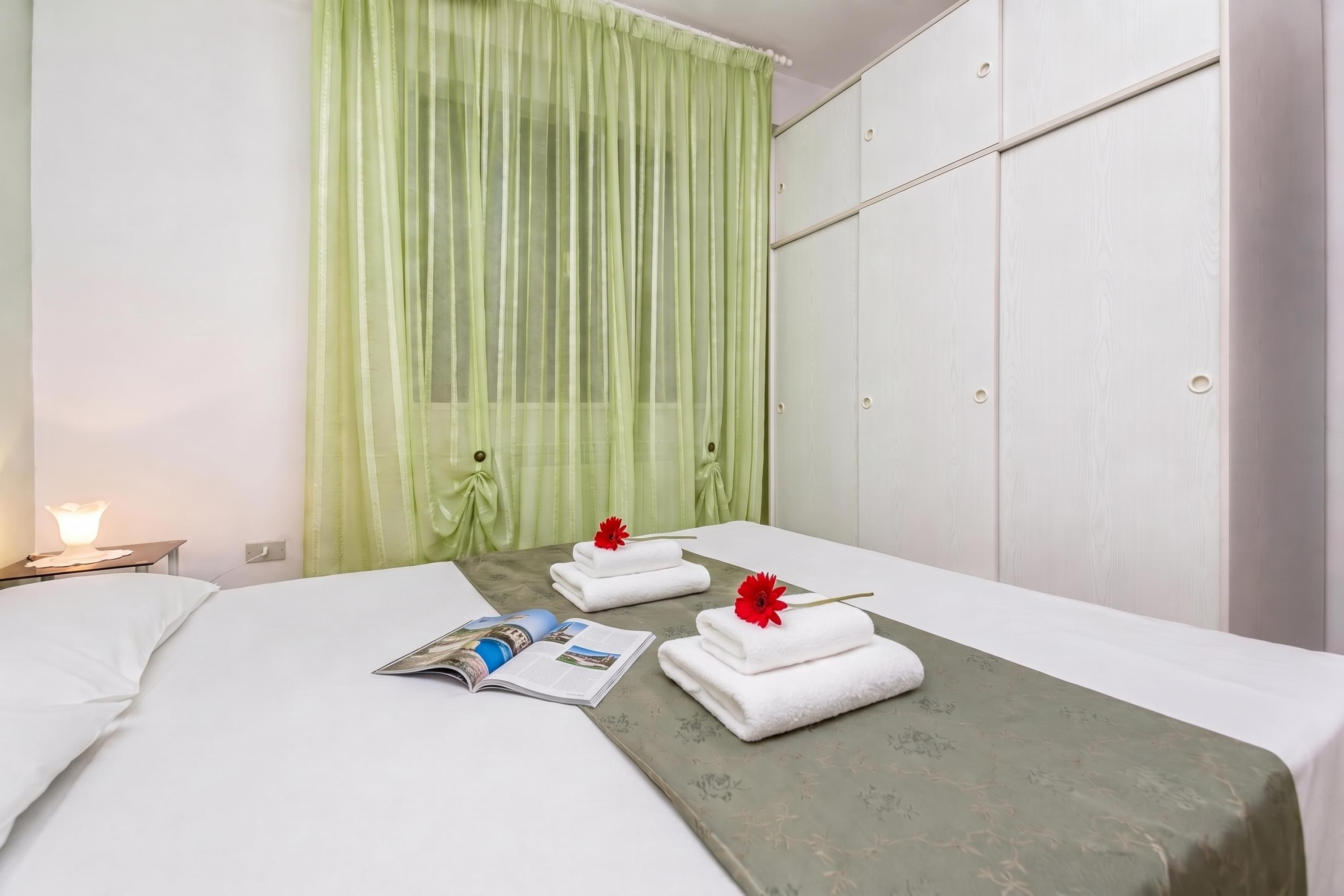 Une chambre d’hôtel avec un lit, une table de chevet avec une lampe, une fenêtre avec des rideaux verts et une serviette ornée d’une fleur rouge.
