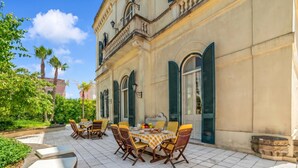 Un área de comedor al aire libre con mantel a cuadros, sillas y vista a un edificio con ventanas de arco y contraventanas.