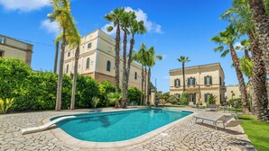 Una piscina con baldosas de piedra, palmeras y un edificio con ventanas arqueadas.
